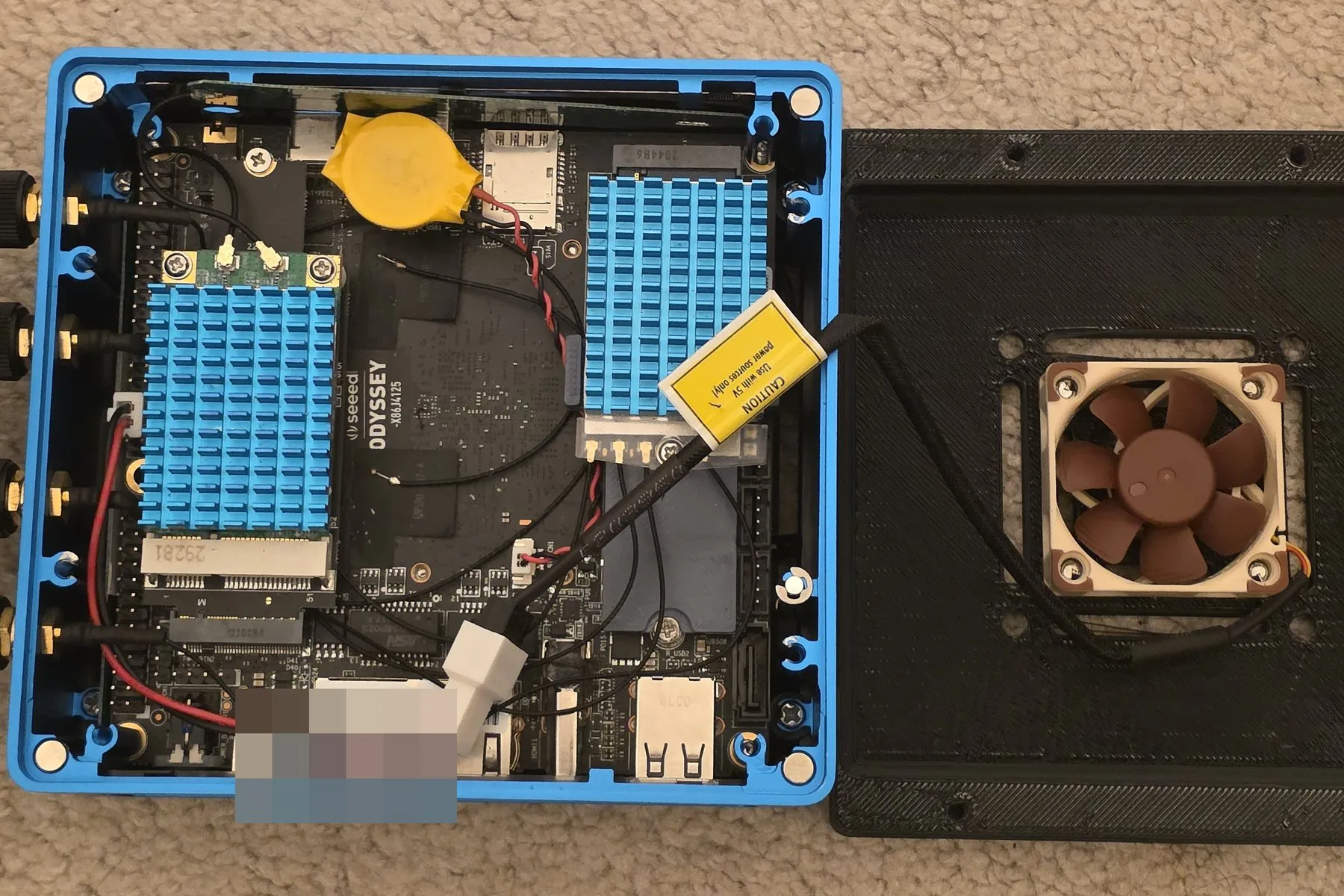 The Seeed Odyssey x86 board opened up: two add-in radio modules under blue heatsinks — the cellular modem and a WiFi card — next to the SIM and microSD slots, with a 3D-printed lid carrying a Noctua fan for cooling.
